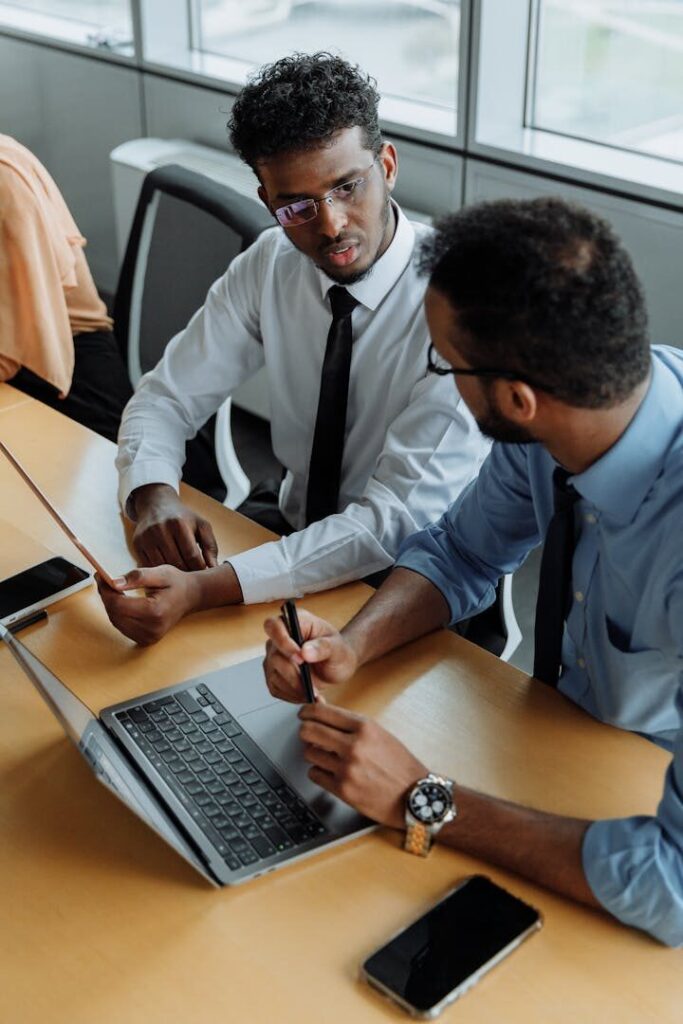 Two men in business attire discuss work at an office desk with a laptop.